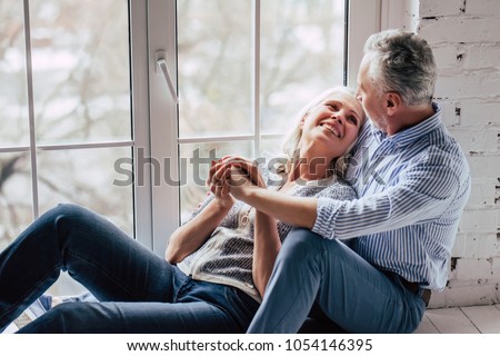Love lives forever! Senior couple at home. Handsome old man and attractive old woman are hugging and enjoying spending time together while sitting on a window sill. Royalty-Free Stock Photo #1054146395