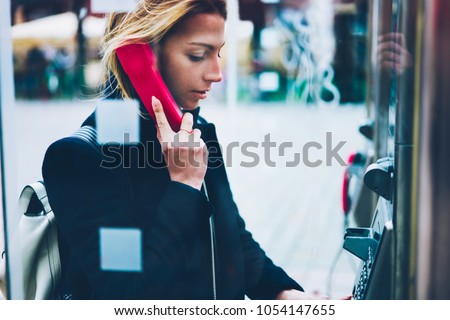 Thoughtful female tourist making call in payphone on street standing in transparent booth, young woman using public telephone operated with coins during travel for call with low prices abroad Royalty-Free Stock Photo #1054147655