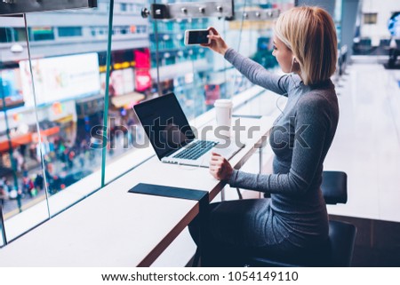 Stylish blonde student making selfie on front camera of modern smartphone for publication on website during listening music online in modern earphones connected to digital laptop device in cafe