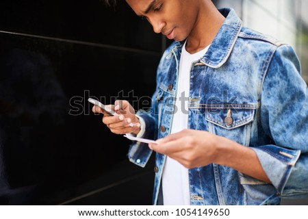Cropped image of pensive hindu hipster guy dressed in denim jacket dialing number on smartphone from business card standing outdoors.Serious blogger checking mail on cellular during walk on street