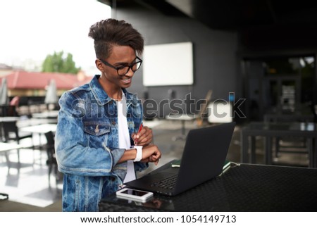 Cheerful hindu student with modern smartwatch looking at laptop computer and checking time on website during remote work outdoors in cafe interior.Positive blogger tracking gps on wristwatch