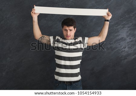 Young tattooed man with blank white banner. Mad man holding advertising sheet, gray studio background, copy space