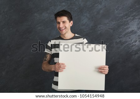 Young tattooed man with blank white banner. Smiling man holding advertising sheet, gray studio background, copy space