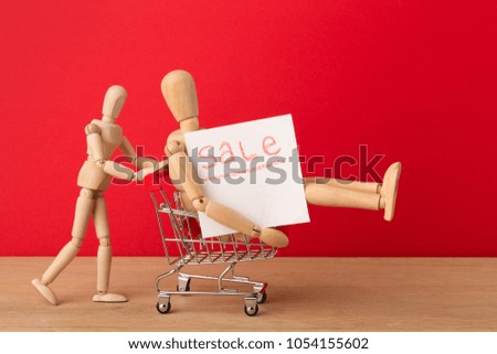 Wooden mannequins with miniature shopping cart running for sales on red background. Mockup for shopping, discount, black friday, copy space, side view.