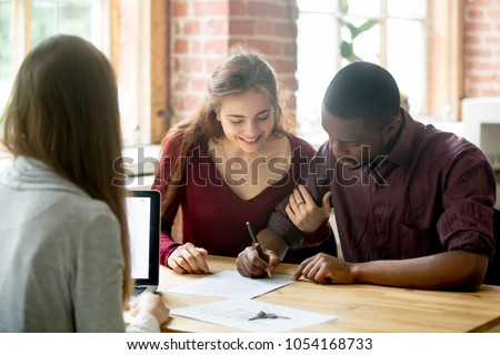 Smiling wife watching african american husband signing purchase deal from female real estate agent. Multiracial diverse couple buying first home together, making loan, legalizing mortgage.