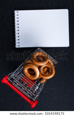 white notebook (mock up design) workup space, shopping list  shopping cart, bagel, corn on dark black background