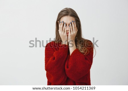 Promise, I am not peeking. Shot of tired and fed up attractive woman in red loose sweater, covering face with palms, feeling stressed and exhausted, having need to relax and sleep over gray wall