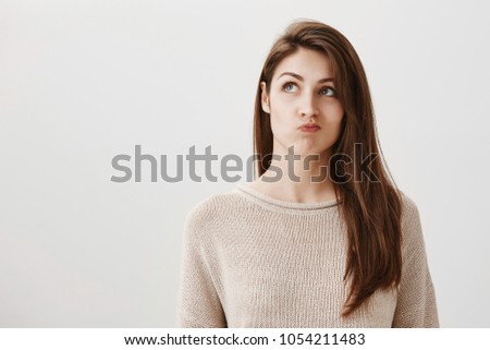 Student is bored making up ideas what to do. Portrait of carefree dreamy caucasian girl looking up with puckered lips, thinking how to entertain herself, wanting to call friend over gray background