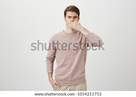 Indoor shot of perplexed and serious european guy covering mouth with hand while holding another in pocket and frowning at camera over gray background. Man promised not to tell a single word