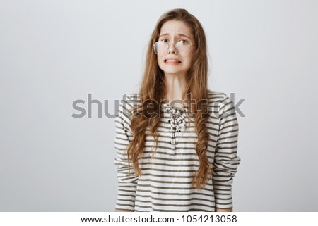 Yikes, bad thing happened. Portrait of confused nervous caucasian teenager in glasses, frowning, clenching teeth and grimacing, feeling embarrassed and awkward, being ashamed over gray wall Royalty-Free Stock Photo #1054213058