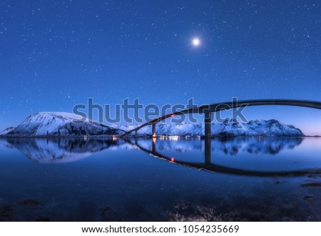 Amazing bridge and starry sky with beautiful reflection in water. Night landscape with bridge, snowy mountains, blue sky with moon and bright stars reflected in sea. Winter in Lofoten islands, Norway 