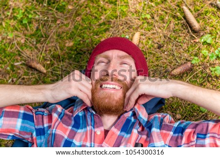 hipster strokes his red beard, a portrait of a curtain, a man lies on the ground in the woods
