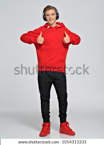 Teenage boy dressed in a red hoodie and street sneakers. Teenager wears fashionable fall clothes and shows thumbs up - posing over white background. Full portrait.