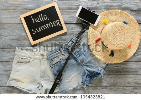 Womens clothing, accessories (denim shorts, straw hat, cellphone, selfie stick, chalkdoard) on grey wooden background. Text Hello summer. Trendy fashion outfit. Shopping, travel concept.  Flat lay
