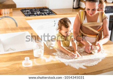 Mother with daughter in sling prepating dough at the kitchen. Concept of food preparation, white kitchen on background. Casual lifestyle photo series in real life interior