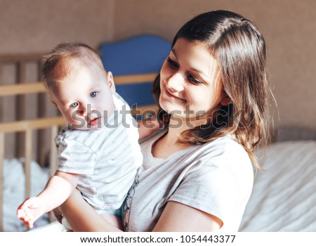 Portrait of adorable blond blue-eyed baby girl looking at the camera with happy smile sitting on his mother's laps in pajamas. Young beautiful woman spending morning with her little doughter