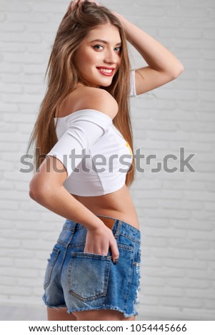 Beautiful Caucasian model in denim shorts and white crop top playfully posing with hand in back pocket and other touching her hair in white photo studio. Blurred brick wall background.