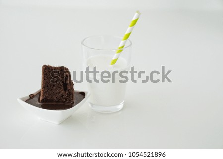Slice of delicious homemade chocolate brownie served on white plate. Served with glass of milk with green white stripe straw. Taken on a white background.