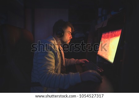 A cute male gamer sits in a cozy room behind a computer and plays games. Gamer plays horror game at night. Computer games. Gamer concept Royalty-Free Stock Photo #1054527317