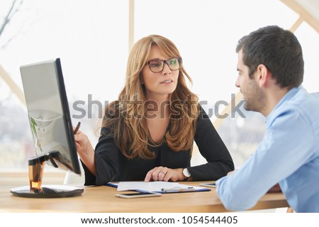 Executive middle aged businesswoman sitting at desk in front of laptop and doing some paperwork while giving sales advice to her young assistant. Brainstorming at the office. Royalty-Free Stock Photo #1054544405