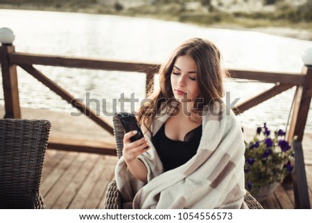 Cheerful relaxed woman uses smart phone for chat with friends, sits in modern coffee shop or terrace cafe. Pretty female reads good news on internet website, enjoys summer rest.
