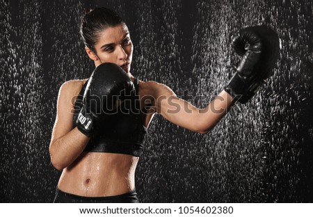 Feminine woman fighter 20s in sportswear doing sports exercises or practising in black boxing gloves while throwing punches under rain drops isolated over dark background