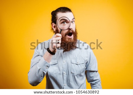 Close-up portrait of cheerful bearded man in shirt looking at camera through magnifying glass, isolated over yellow background Royalty-Free Stock Photo #1054630892