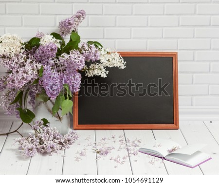 Beautiful bouquet of white and violet lilac flowers in glass vase with opened pad and black chalkboard,on the white wooden and brick background with empty place for text, mock up front view