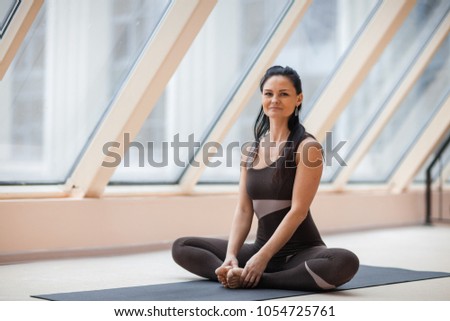 Front view portrait of beautiful young woman working out against large windows, resting after doing yoga exercises, sitting in ardha Padmasana, Lotus pose, relaxing. Full length