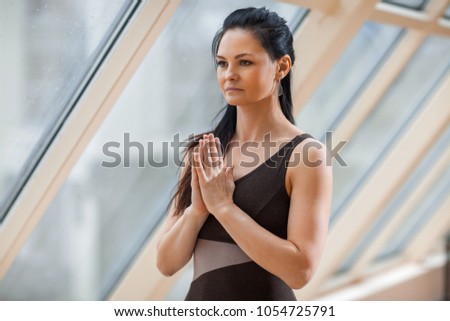 portrait of sport a smiling attractive woman brunette in a black jumpsuit making the Namaste gesture, practicing yoga, working out, wearing sportswear, large windows background