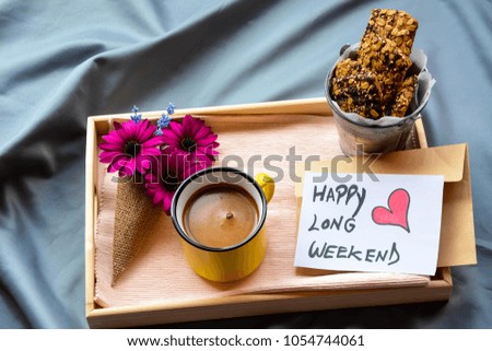 A wooden tray on the bed with flowers ,coffee ,Granola bars and Happy Long Weekend text on a note