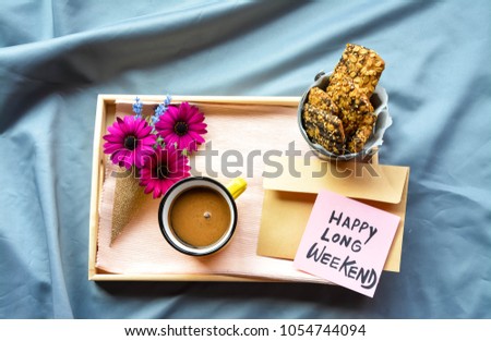 A wooden tray on the bed with flowers ,coffee ,Granola bars and Happy Long Weekend text on a note