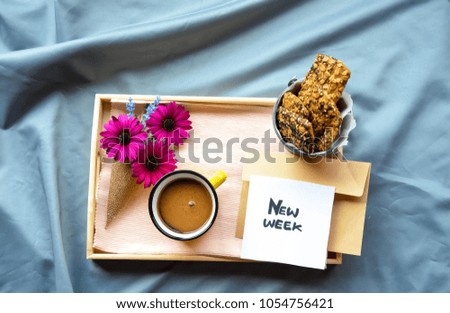 A wooden tray on the bed with flowers ,coffee ,Granola bars and new week text on a note