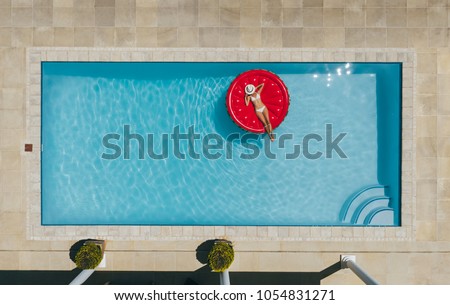 Aerial view of female in bikini lying on a floating mattress in swimming pool with her face covered with hat. Top view of woman sunbathing on inflatable mattress in pool.
