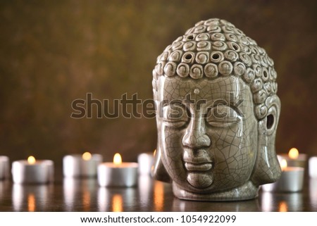 Buddha head statue standing on a dark stone surface, burning candles all around it, meditation or relax spa treatment concept, front view