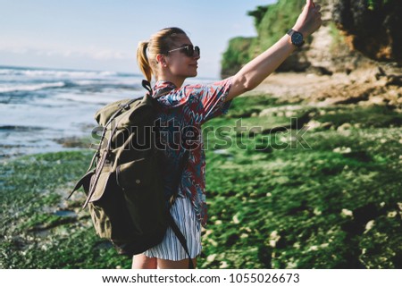 Positive young hiker with rucksack exploring environment of green island with high hills and making selfies for publicating in travel blog.Cheerful female tourist taking photos during summer trip