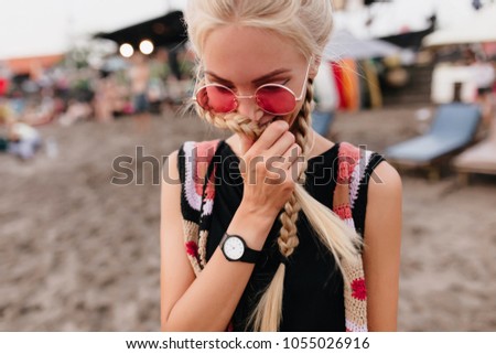 Shy blonde woman with braids posing at beach. Outdoor portrait of cute fair-haired girl in pink sunglasses and black tank-top.