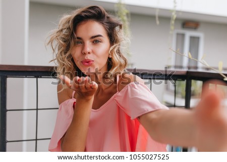 Glamorous blonde woman in pink attire making selfie while sending air kiss. Attractive female model with wavy hairstyle taking picture of herself at balcony.