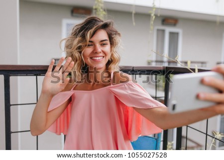 Cheerful tanned girl using smartphone for selfie at balcony. Magnificent blonde lady in pink attire taking picture of herself and waving hand.