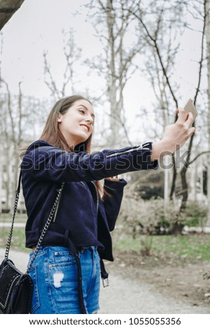 beautiful brunette woman wearing a suede leather jacket, ripped jeans and a black purse. attractive fashion blogger posing outdoors, smiling in a park, taking selfie photo with her smartphone.