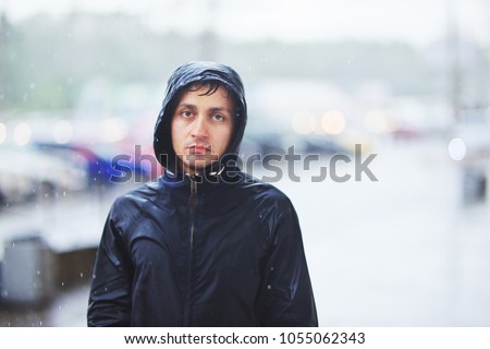 Portrait of a sad man in a raincoat and a hood in the rain. Bad stormy rainy weather sorrow concept. Royalty-Free Stock Photo #1055062343