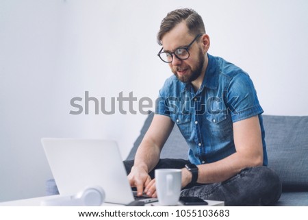 Young businessman typing on his laptop sitting on a sofa in cozy co-working. Distant job concept. Empty white wall on the background. Copy space for your text.