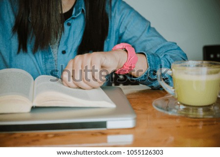 Hands of young woman looking at watch while reading a book on wooden table.