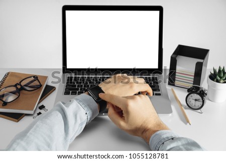 View of male hands checking his smart watch in front of a  laptop with blank white screen standing on the white office desk. Mock up, copy space for your text.