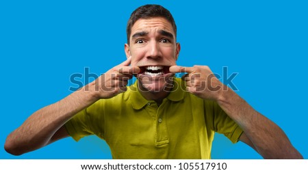 Handsome young man doing a funny pose pulling the corner of his mouth with his fingers. Over blue editable monochrome background
