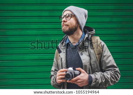 Male casually dressed bearded tourist with a backpack posing with retro film photo camera in front of bright green roller shutter. Traveling and freelance concept.