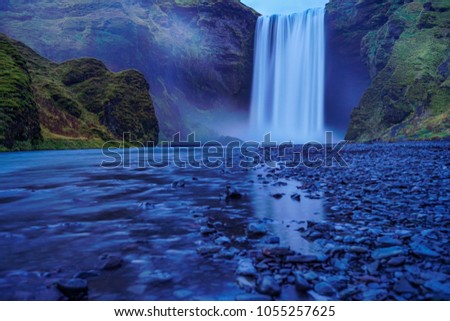 Long exposure photography Skogafoss waterfall during late evening under blue cloudy sky with rocks in foreground , Iceland                              