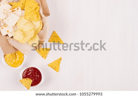 Golden different snacks on craft paper, triangles nachos, red and yellow sauce in bowl on soft white wood background, with copy space, border. Summer fresh fast food background.