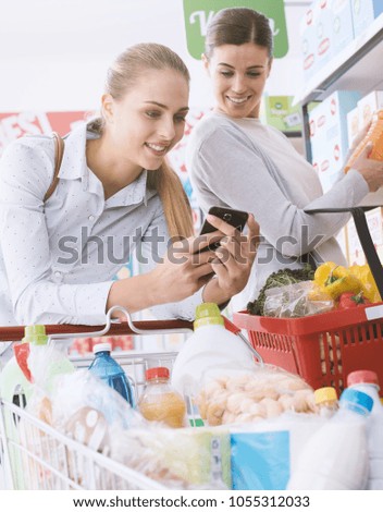 Happy female friends shopping together at the supermarket, they are searching products and offers using apps on their smartphone
