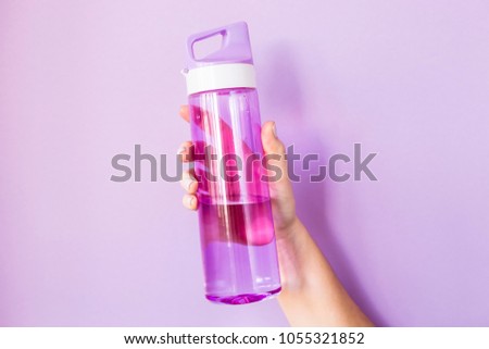 A woman holds a purple water bottle in her hand for sports.  On a bright purple background. Healthy lifestyle and fitness concept Royalty-Free Stock Photo #1055321852